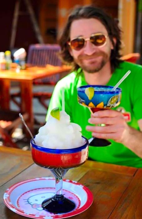 A large frozen margarita placed on a wooden table, with Randy holding up a margarita in the background