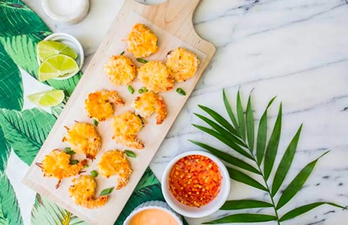 Overhead shot of coconut shrimp on a wooden tray surrounded by dipping sauces and limes.