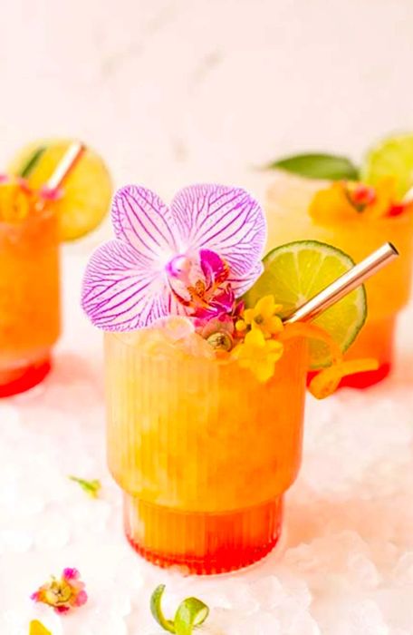 Pineapple mango rum punch cocktail adorned with a large pink flower, a lime slice, and a twist of orange zest