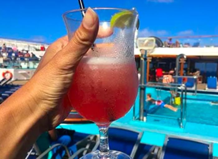 A hand holding a pink margarita with a pool visible in the background on the Dinogo Conquest.