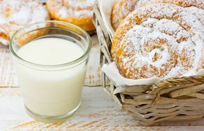 A plate of four swirl-shaped pastries, known as ensaimada, dusted with powdered sugar, accompanied by a glass of milk.