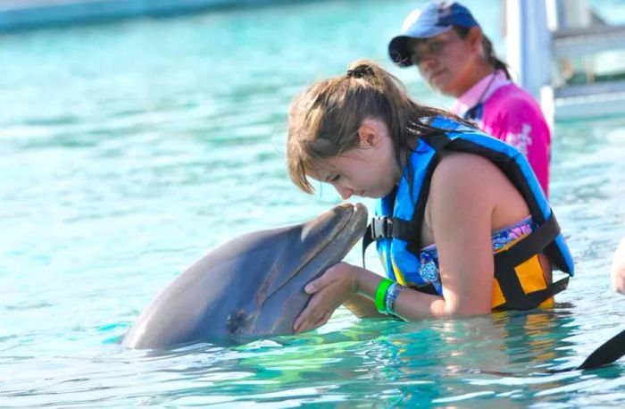 girl kissing a dolphin while a trainer supervises