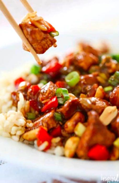 A pair of chopsticks lifting a piece of chicken from a flavorful Kung Pao Chicken dish.