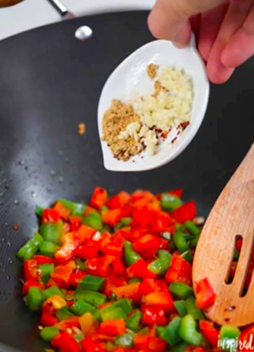 A hand holding a small bowl of minced garlic and ginger above red and green bell peppers with red pepper flakes in a skillet