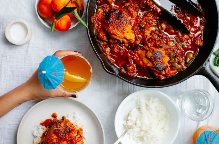 A plate of pollo guisado and rice on a table with a hand reaching for a citrus rum spritzer