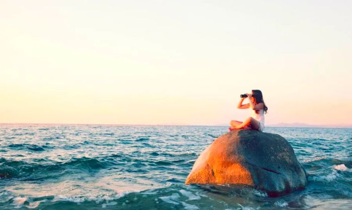 A child perched on a seaside rock, scanning the horizon with binoculars.