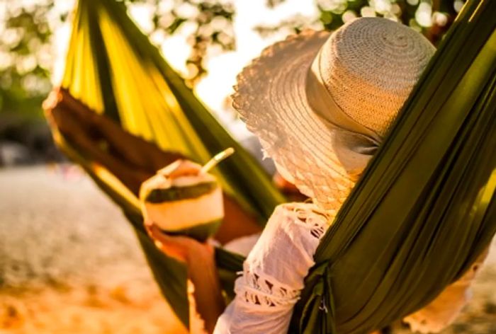 A woman relaxes in a hammock in the Caribbean while sipping a coconut cocktail.