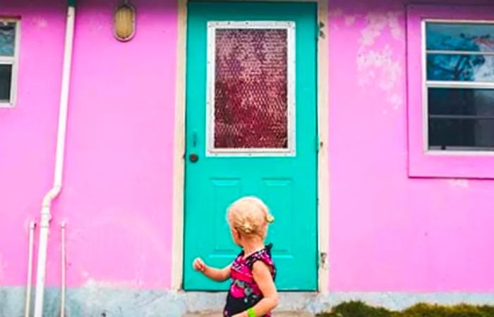 A young girl standing in front of a pink building with a green door