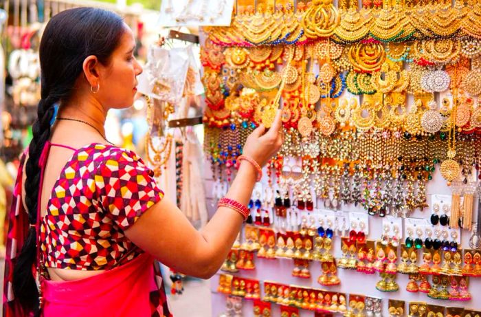 An Indian woman in her mature years is browsing and purchasing earrings and jewelry at an outdoor street market in Delhi, India, during the daytime. She is dressed in a traditional Indian sari.