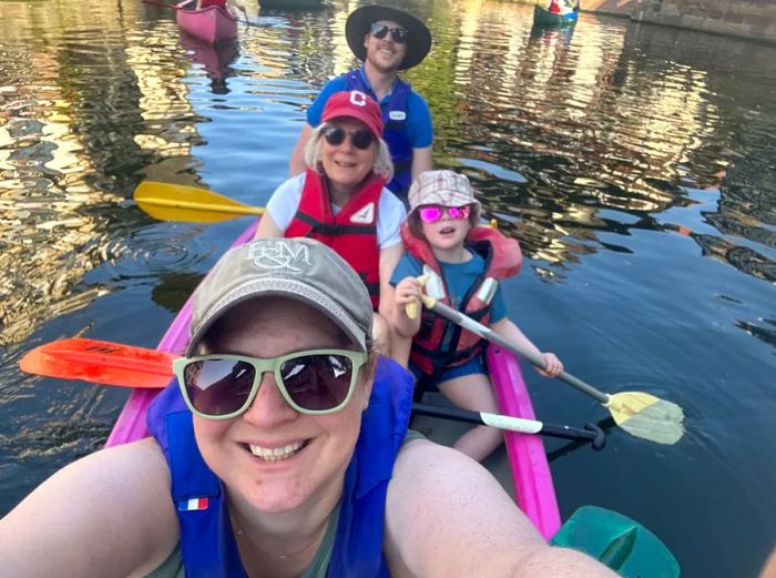 A multigenerational family of four smiles for a selfie in a pink canoe, holding paddles while wearing hats and sunglasses.