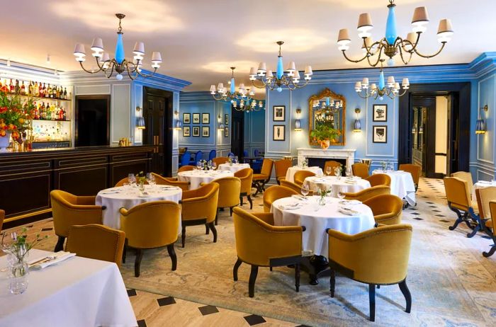 The dining area features a bar to the left, light blue walls, simple chandeliers, and yellow velvet dining chairs arranged around circular tables for four.