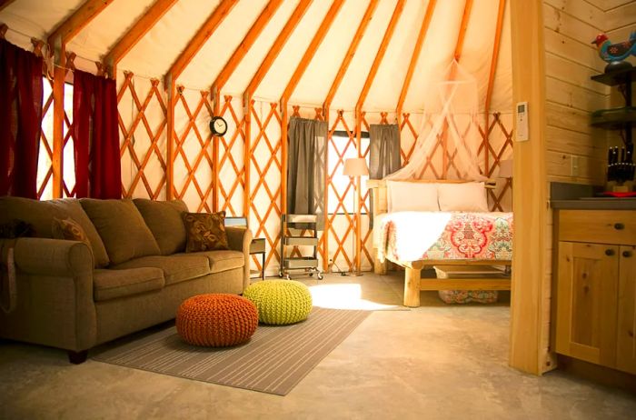 Inside a yurt, featuring a bed, sofa, and the edge of a light-wood kitchen to the right.