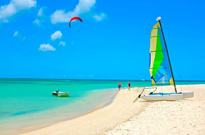 A stunning Caribbean beach featuring turquoise waters, soft white sand, a vibrant red parasail, and a small sailboat adorned with blue, green, and yellow sails.