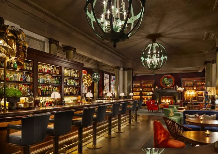 A dimly lit bar with a cozy fireplace in the backdrop, showcasing geometric light fixtures and a row of stools lining a lengthy bar.