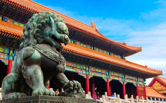 In the foreground stands a metal lion statue, with a palace building of the Forbidden City featuring ancient Chinese architectural elements in red and gold hues behind it.