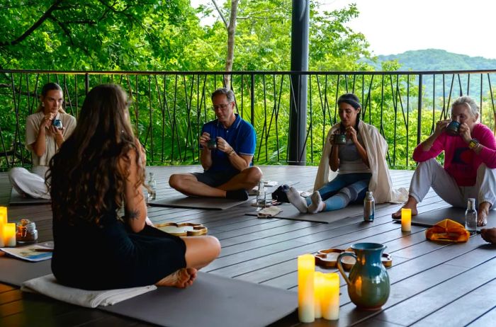 A few students are seated on the wooden floor of an open-air pavilion, participating in a cacao ceremony, surrounded by several flickering candles.