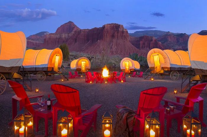 At dusk, red Adirondack chairs and lanterns encircle six lit Conestoga wagons, with rugged mountains rising in the distance.