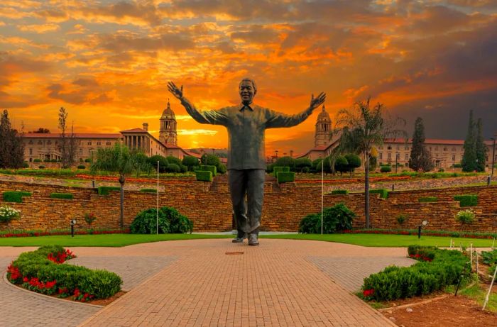 A prominent statue of Nelson Mandela stands with arms wide open in front of the historic Union Buildings in Pretoria, South Africa, surrounded by well-maintained landscaping.