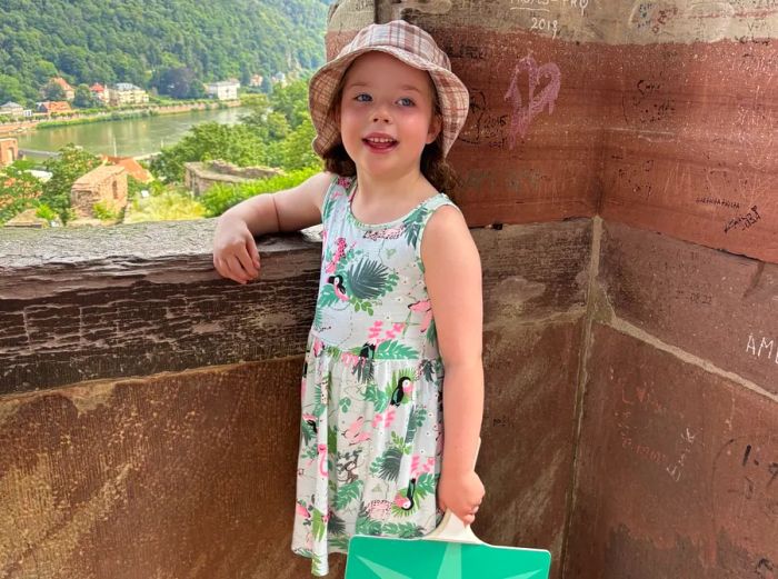 Fran Golden's six-year-old granddaughter poses by a castle window overlooking the Rhine River, sporting a bucket hat and a dress while holding a green sign.