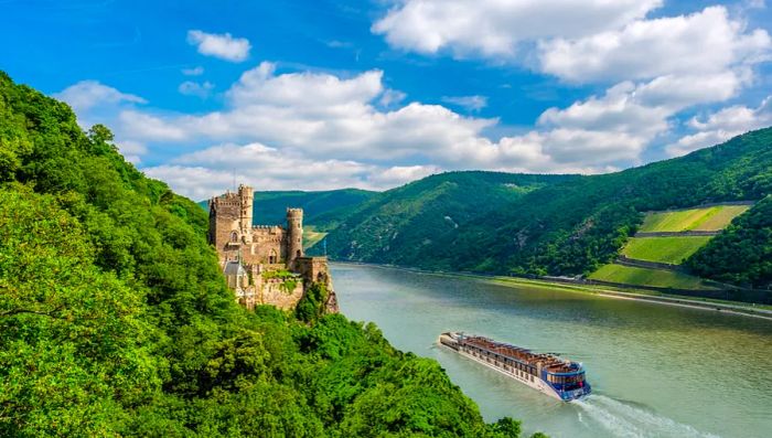 An AmaWaterways river cruise ship gliding through the Rhine River Gorge, passing by a castle perched on a verdant hillside.