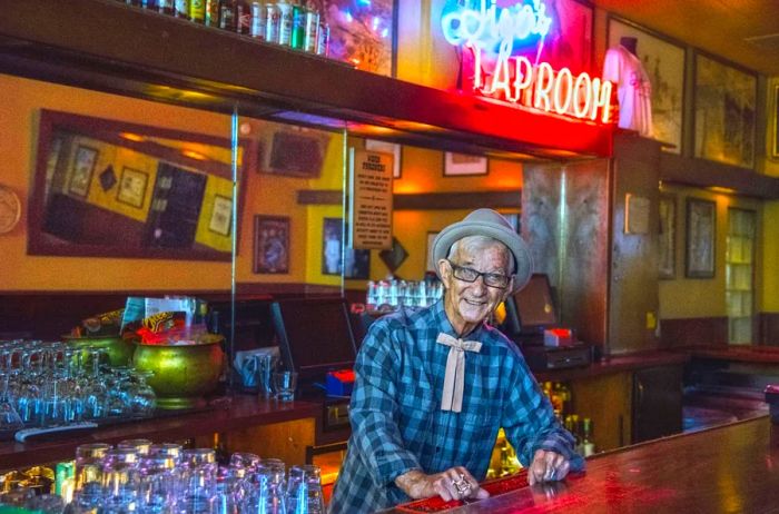 An elderly man in a checkered shirt and hat stands behind a bar illuminated by a neon sign.