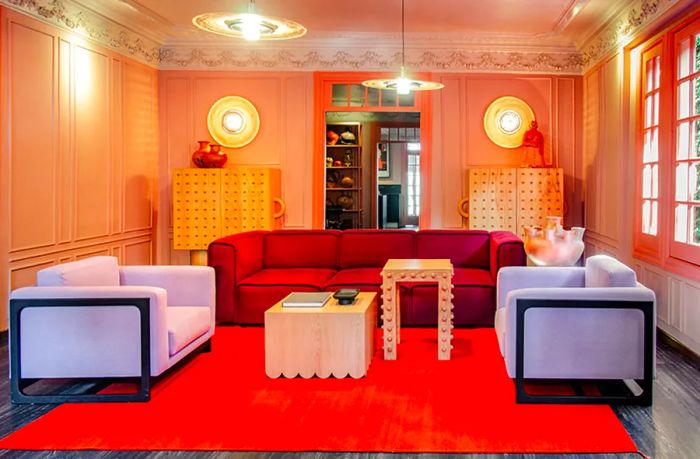 A bright, stylish sitting room at Ignacia Guest House in Mexico City, featuring a red square couch, white chairs, and an orange rug.