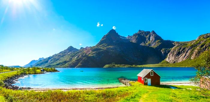 A serene bay with shallow waters, featuring a small red house by the shore and mountains in the background, illuminated brightly at 9 p.m. in a Norwegian summer.