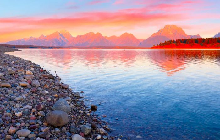 The sun rises over Jackson Lake, with rocky shores to the left and rugged mountains in the distance