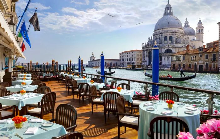 A terrace featuring clothed tables beside a canal in Venice, framed by a stunning cathedral in the distance.