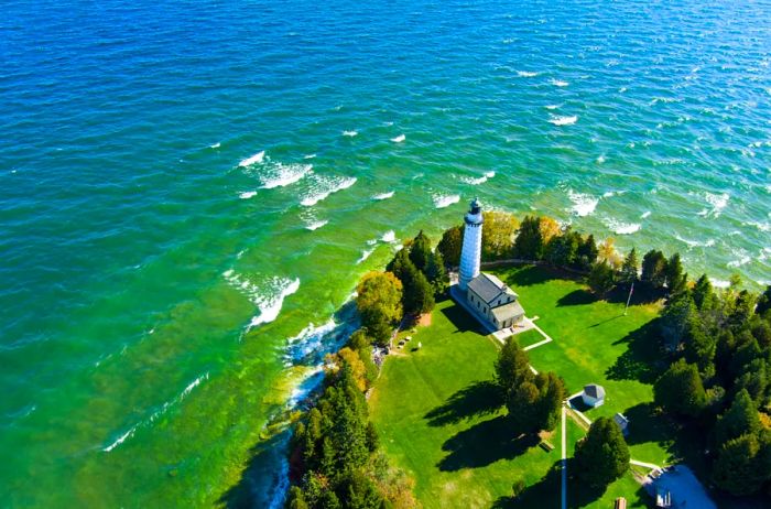 Aerial view showcasing a white lighthouse perched on lush green land, surrounded by shimmering blue-green waters.