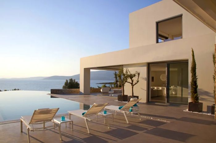 Exterior view of a modern beige villa in Greece featuring an infinity pool and multiple deck chairs.