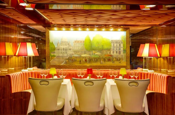 A dining setup in the restaurant aboard Uniworld's 'S.S. Joie de Vivre', featuring a banquette, dining chairs, and a painting of the Sacré-Cœur adorning the wall, complemented by striped table lamps.