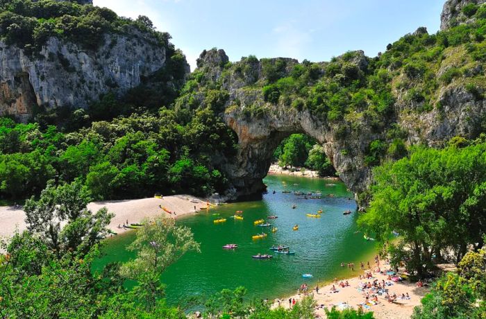 A vibrant array of kayaks rests in the water beneath the Gorges de l’Ardèche in France, with sunbathers relaxing on the sandy shore nearby.