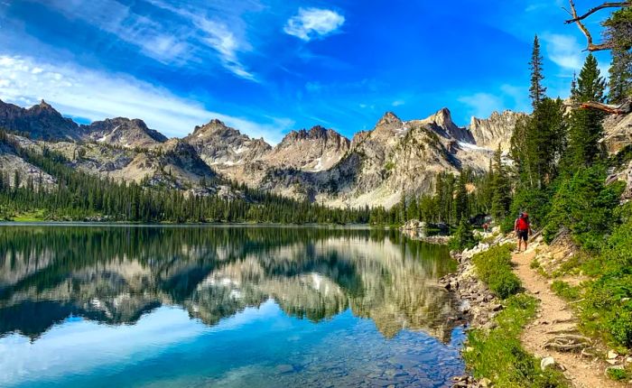 A serene lake nestled among mountains and pine trees, their reflections shimmering on the water's surface, features a male hiker with a red backpack along the trail.