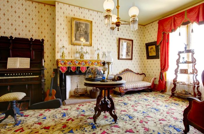 An interior of a historic 1857 house showcasing a patterned carpet, decorative wallpaper, a grand wooden piano, and a striking black marble fireplace.