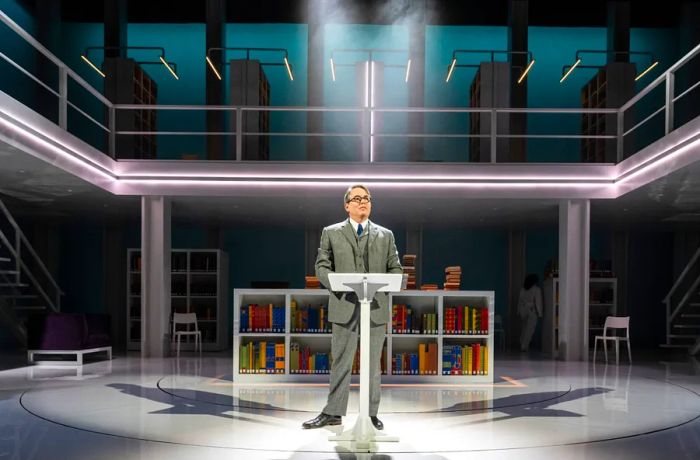 A man in a suit stands at a podium on stage, with a bookshelf and a lit balcony behind him.