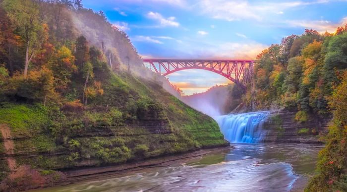 A river and waterfall cascade through the striking limestone rock canyon of Letchworth State Park, with a soaring red bridge in the background framed by trees showcasing their fall colors.