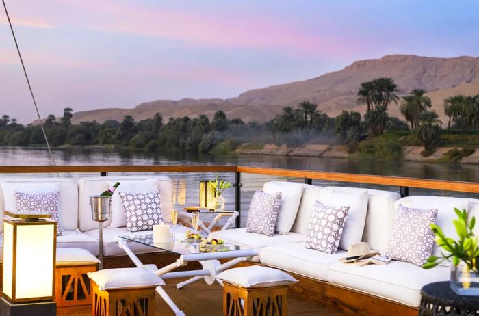 The rooftop deck of the Sanctuary Zein Nile Chateau features white outdoor couches, lanterns, and champagne, all set against the stunning backdrop of the Nile River desert and palm trees.