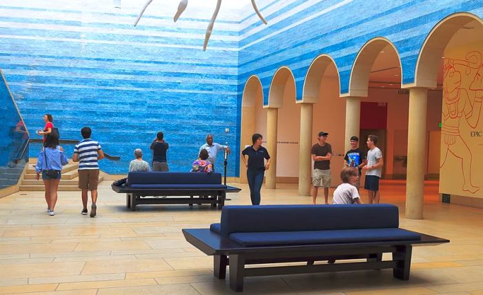 Individuals seated and strolling in a foyer adorned with blue walls, benches, and an arched loggia.