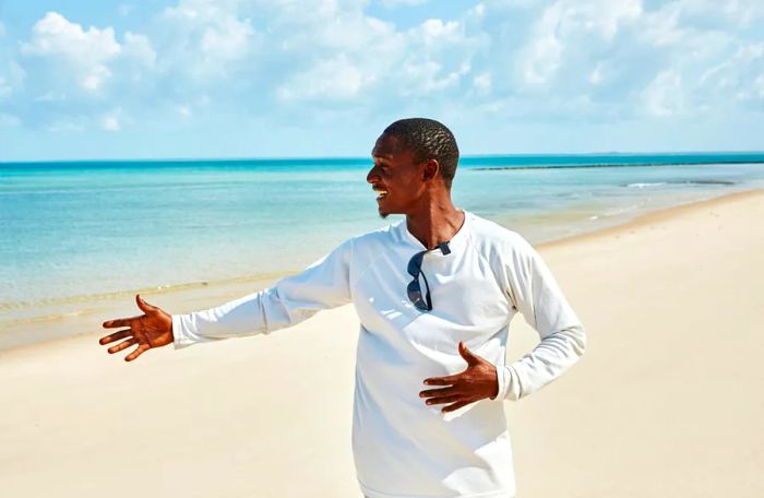 A man wearing a white long-sleeve shirt extends his right arm while standing on the beach.