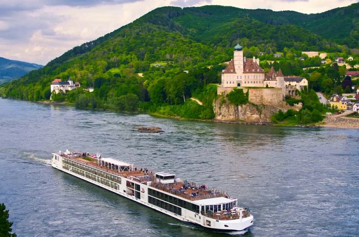 A Viking Cruises longship gliding along the Danube River, with a historic castle and lush hills in the backdrop.