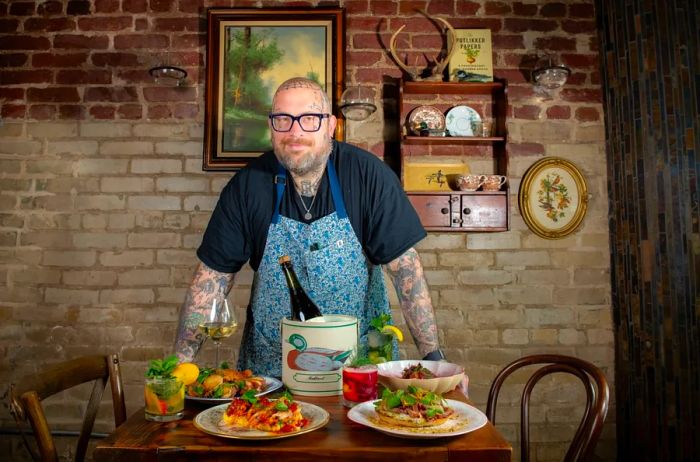 A chef, sporting a tattooed shaved head and arms, stands behind a table laden with food plates and a wine bucket adorned with a duck.