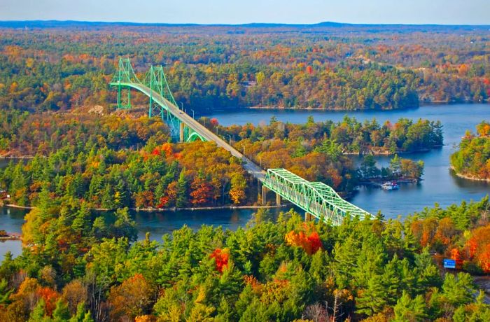 An aerial perspective showcases a long, mint-hued road bridge spanning the Thousand Islands–Seaway over the St. Lawrence River, with hints of fall colors emerging among the tree-covered islands.