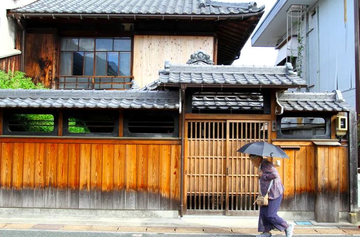 The exterior of an old house in Udatsu, complete with a wooden fence and gray tiled roofs; a person holding a black umbrella is in the foreground.