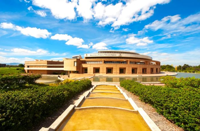 A distant perspective of the curved exterior of the Virgilio Barco Library, featuring a moat and lush green shrubs.