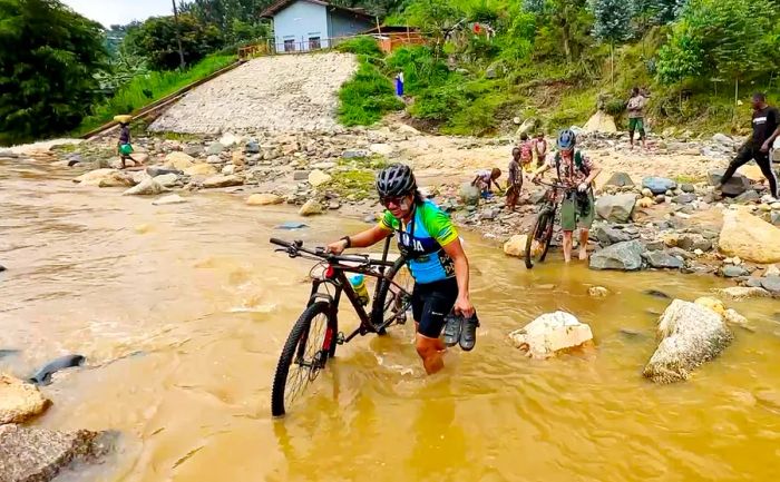 Cyclists wading through a muddy river while carrying their bicycles on foot.