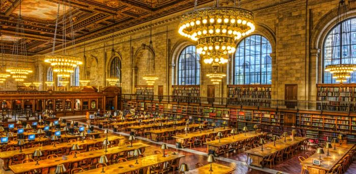 Aerial perspective of the Rose Main Reading Room in the main branch of the New York Public Library located at 5th Avenue and 42nd Street, showcasing large chandeliers above rows of wooden tables illuminated by small lamps, alongside a wall adorned with arched windows.