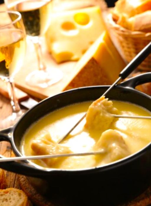 A pot of melted cheese filled with fondue sticks topped with bread, accompanied by two glasses of wine in the backdrop.