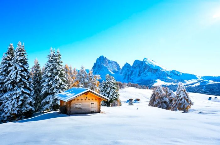 A cozy wooden cabin nestled in a snowy landscape, encircled by mountains and snow-laden pine trees.