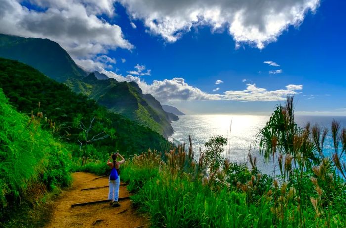 A hiker captures a moment on a dirt trail surrounded by lush tropical vegetation, verdant mountains, and the ocean beyond.